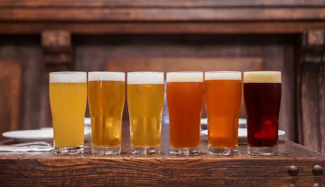Row of six craft beer glasses showcasing different colors and styles, placed on a rustic wooden bar.
